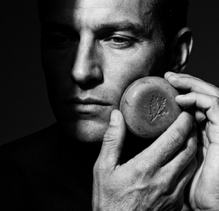 Black and white photo of a man holding a round natural and handmade soap bar close to his face.
