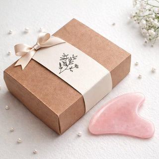 Pink gua sha tool next to a brown gift box with a white ribbon on a light background