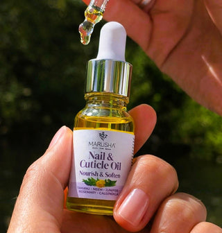 Marusha nail and cuticle oil being held with a dropper above it against a blurred green background.