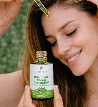 Woman applying hair growth oil to her scalp with a dropper, holding a bottle labeled 'Marusha'.