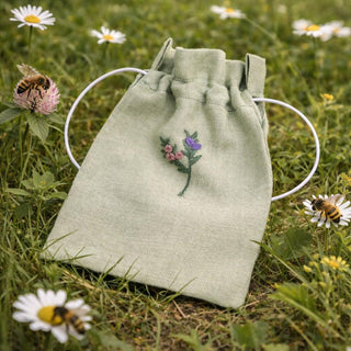 Green drawstring pouch with floral embroidery on grass with flowers and bees
